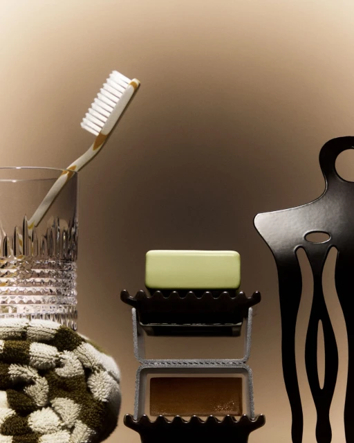 Still life of bathroom items: toothbrush in glass, green bar of soap on holder, dark brown bar of soap, patterned towel, chair.