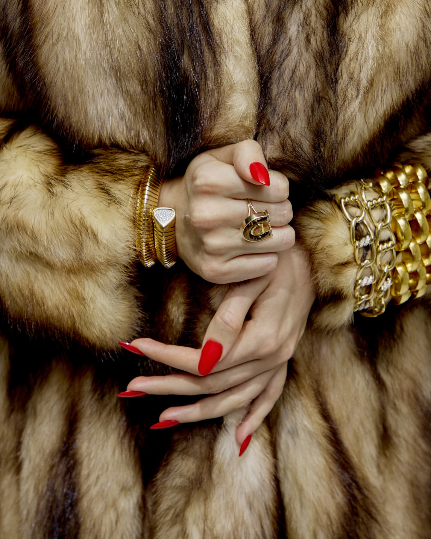 Close-up of hands adorned with gold bracelets and a ring, featuring red manicured nails, against a blurred fur coat background.