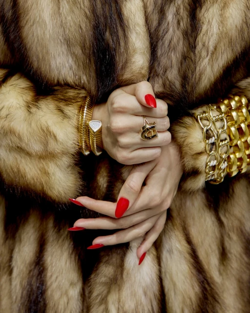 Close-up of hands adorned with gold bracelets and a ring, featuring red manicured nails, against a blurred fur coat background.