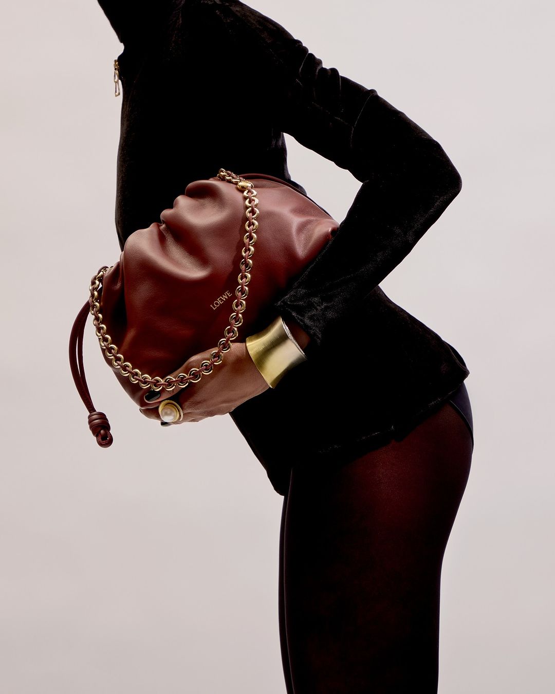 Fashion shot featuring a model holding a red LOEWE bag with a gold chain strap, wearing a black velvet top and gold cuff bracelet.