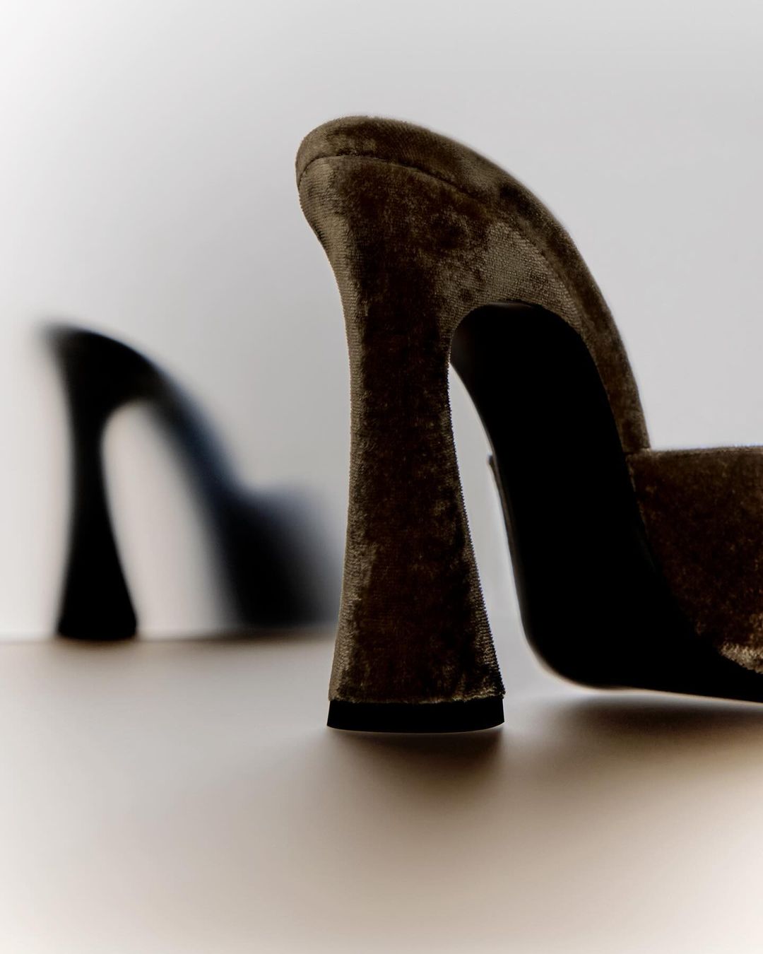 Close-up of olive-green velvet high-heel mule with another pair blurred in the background against a white backdrop.