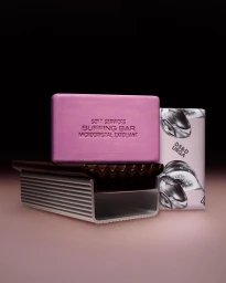 Pink exfoliating body soap bar resting on a ribbed metallic soap dish with a patterned soap box beside it