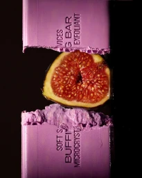 A fresh fig cut in half rests between two broken halves of a lilac exfoliant soap bar against a dark background.