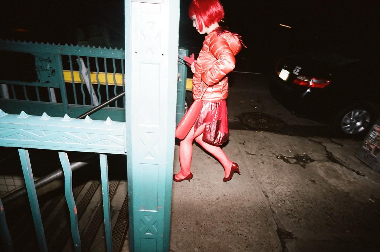 Woman in red wig and outfit walks from subway entrance at night. She wears a puffy jacket, short dress, matching tights and heels.