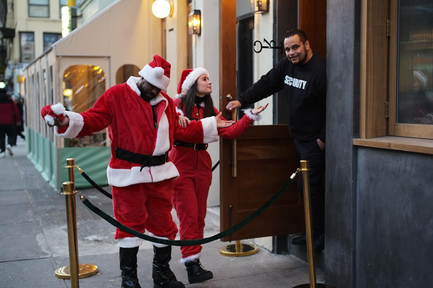 Two individuals in Santa Claus costumes stand outside a building with a security guard standing by the entrance, creating a festive scene.