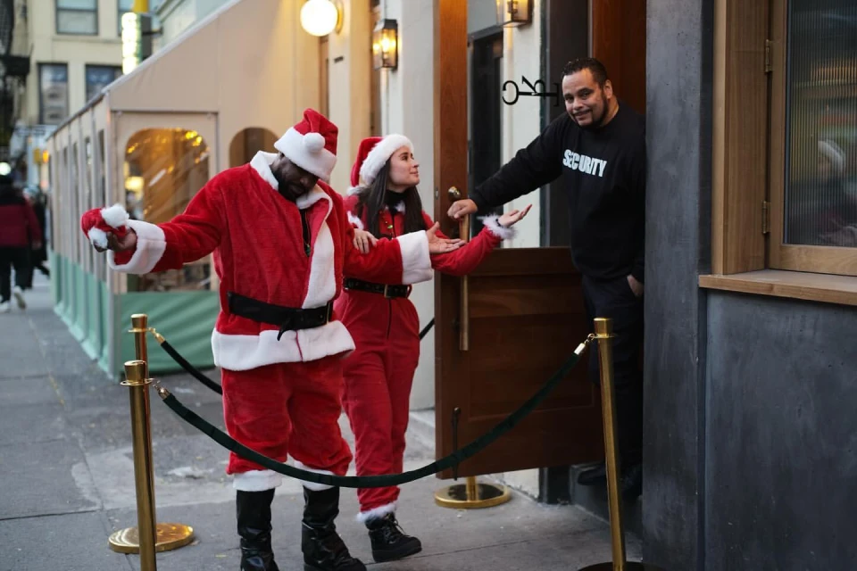 Two individuals in Santa Claus costumes stand outside a building with a security guard standing by the entrance, creating a festive scene.