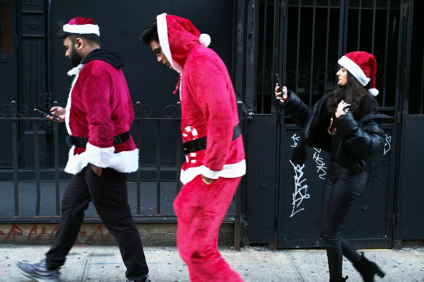 Three people in Santa Claus costumes walk on a sidewalk in front of a black metal fence, one taking a picture with a smartphone.