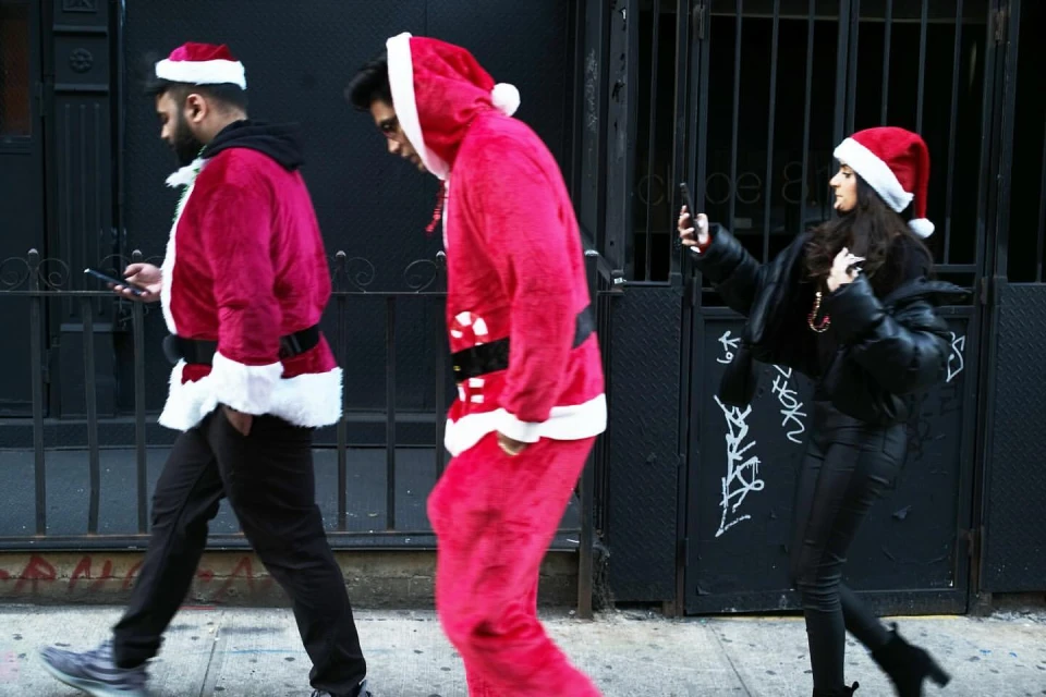 Three people in Santa Claus costumes walk on a sidewalk in front of a black metal fence, one taking a picture with a smartphone.