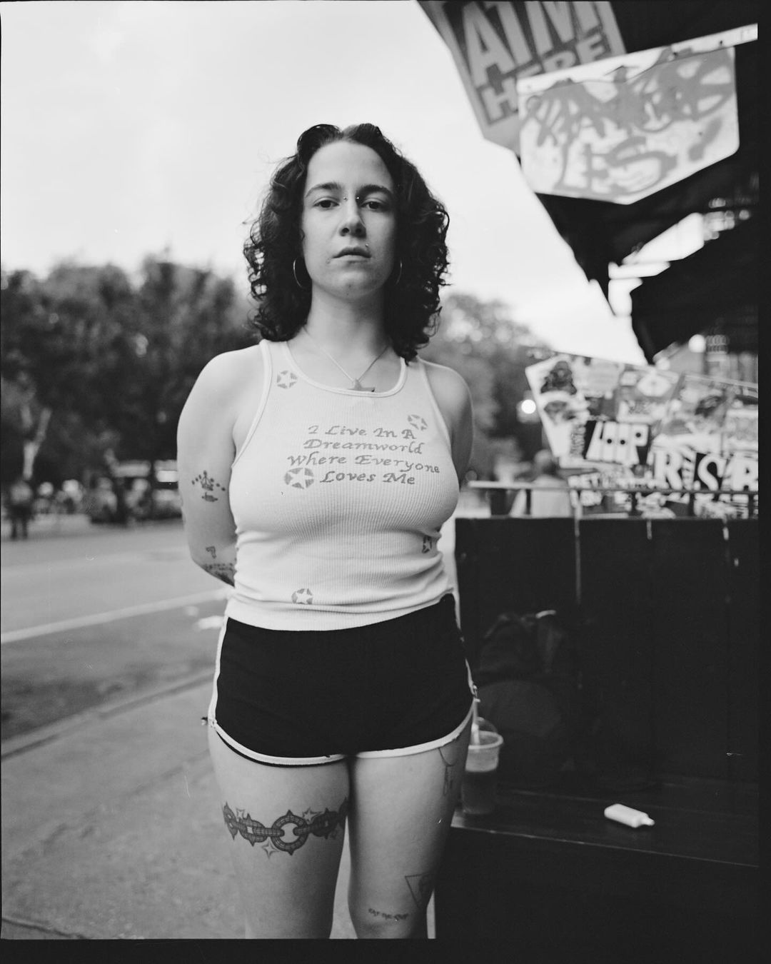Monochrome portrait of a woman with curly hair, wearing a tank top and shorts, standing outside with a neutral expression.
