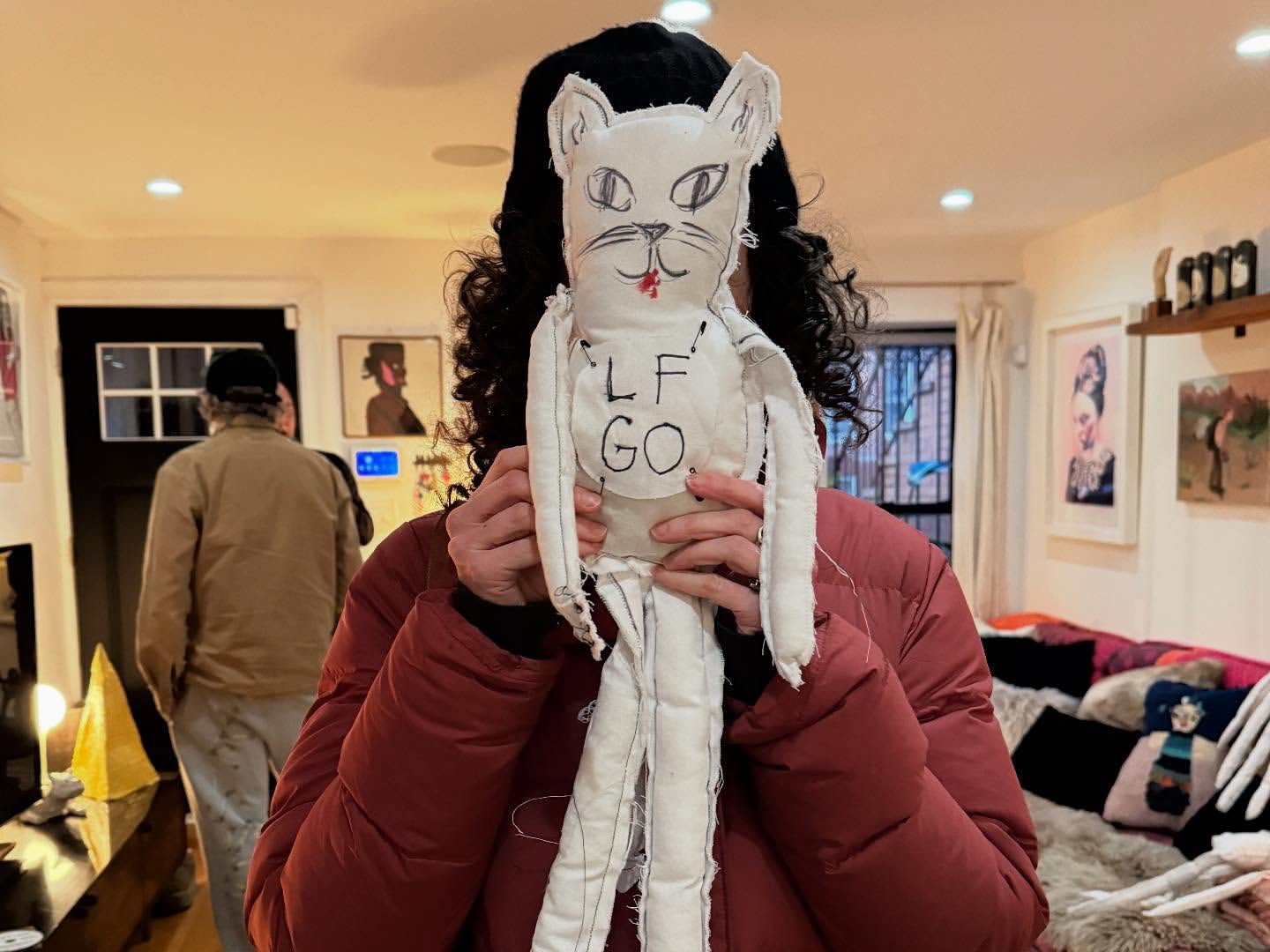 Person in a red puffer jacket holding a white handmade cat doll with 'LF GO' written on it, standing in an indoor setting.