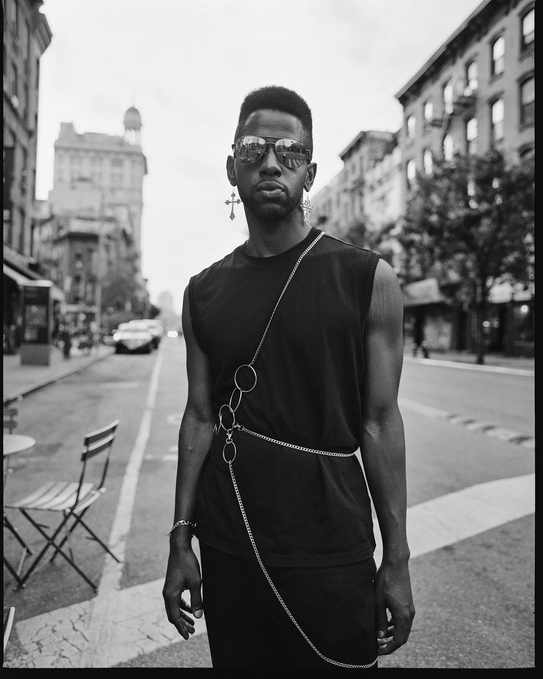 Black and white portrait of a stylish man in sunglasses and a sleeveless shirt, posing on a city street with buildings in the background.