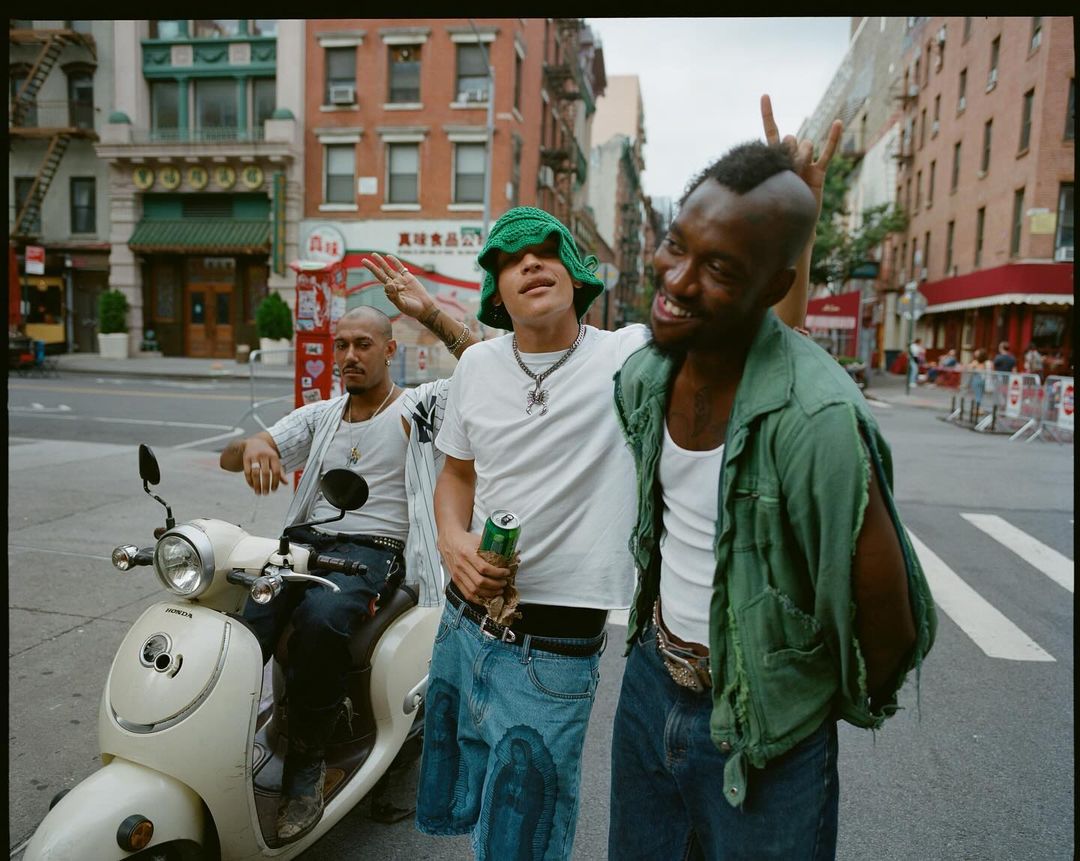 Three young men in casual fashion pose on a city street next to a cream moped, capturing urban youth culture and street style.