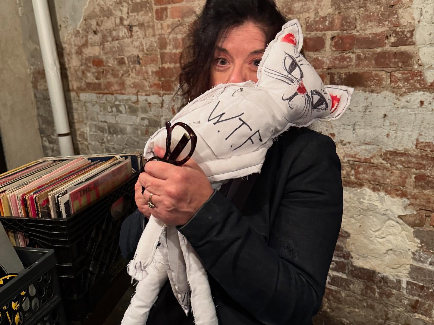 Adult female holds a stuffed white cat with black lettering (WTF) in front of a partially visible brick wall and record collection.