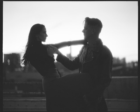 Black and white image of an adult couple interacting on a rooftop against an overcast sky, creating a silhouette effect and a moody atmosphere.