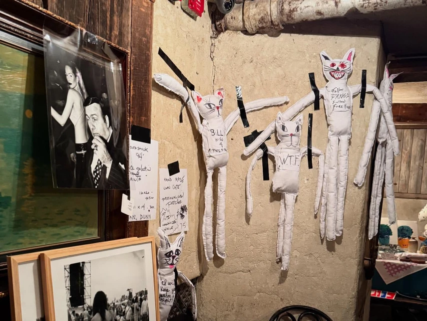 Indoor shot of art-covered walls with framed photos, paintings, and fabric dolls. The room has a gritty, textured aesthetic with eclectic decor.