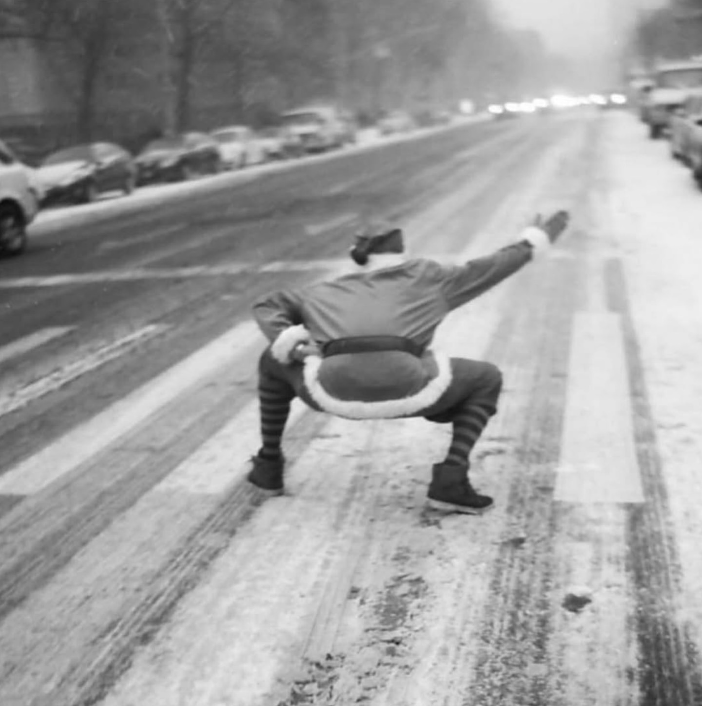 Person dressed as Santa crouches in a crosswalk in snow, creating a humorous, festive image in an urban environment with cars parked on the street.