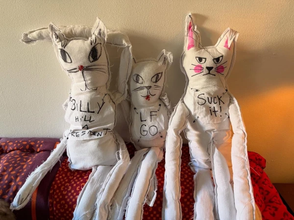 Three handmade white cat dolls with text, arranged on a patterned surface, evoke a whimsical and slightly unsettling aesthetic.