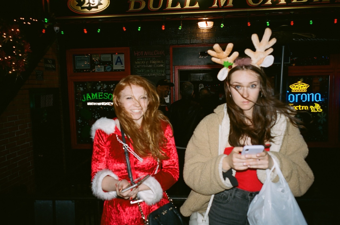 Two women pose outside Bull McCabe's bar; one in a Santa dress and the other in reindeer antlers with a phone.