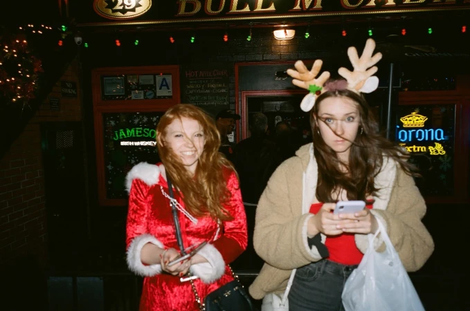 Two women pose outside Bull McCabe's bar; one in a Santa dress and the other in reindeer antlers with a phone.