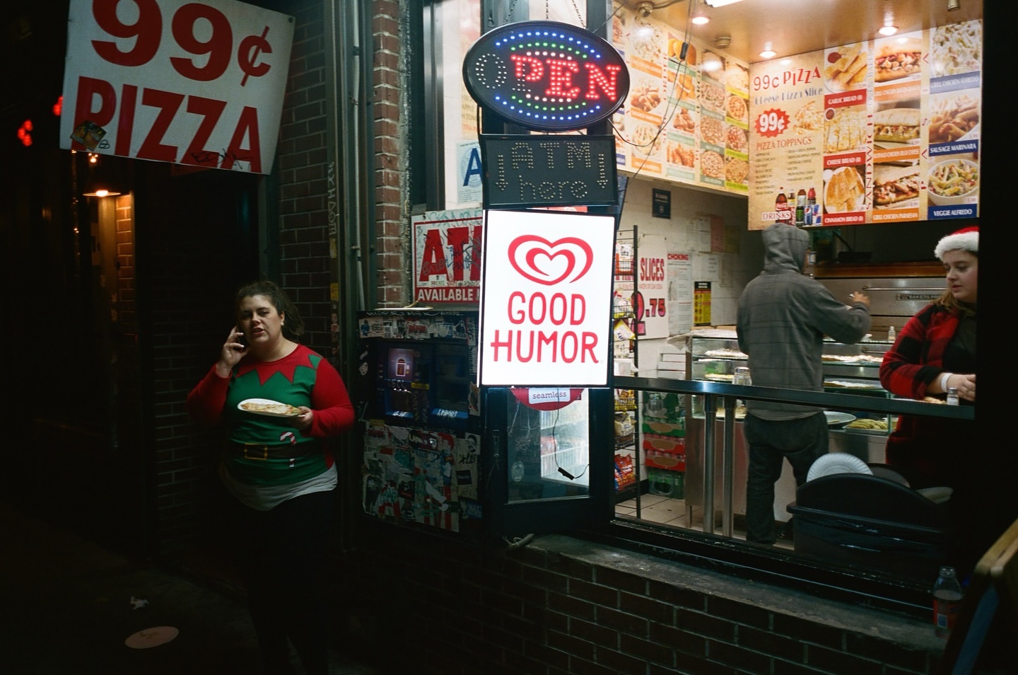 Woman on phone stands outside 99 cent pizza shop at night next to ice cream sign. People inside store visible.