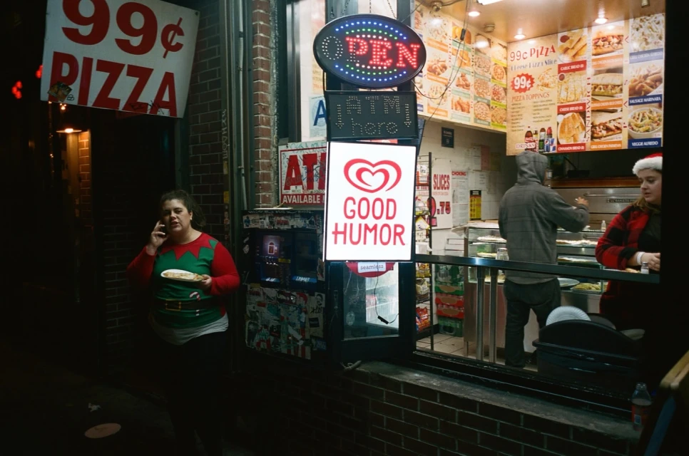 Woman on phone stands outside 99 cent pizza shop at night next to ice cream sign. People inside store visible.