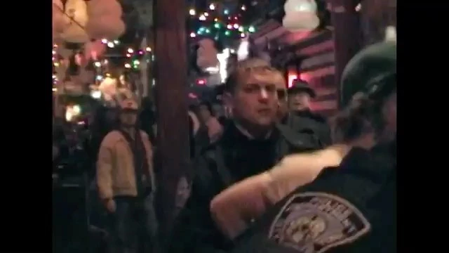 Nighttime crowd scene with a fair-skinned adult male looking forward, surrounded by a group of people and festive string lights.