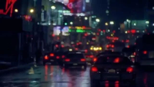 Nighttime city street with traffic, headlights, tail lights, and neon signs reflecting on the wet pavement, creating a vibrant urban scene.