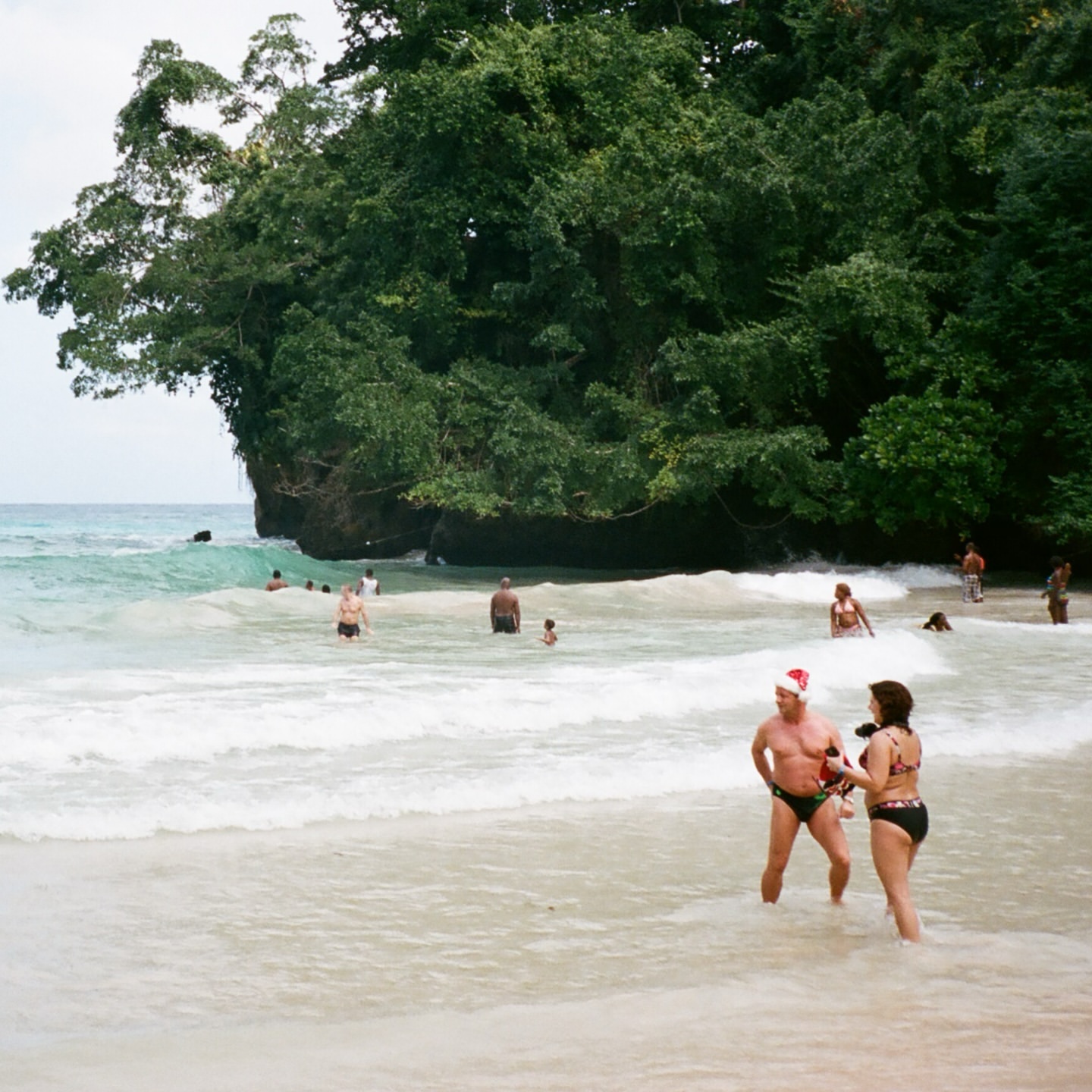 People in swimwear wade in the ocean surf near a beach with dense tropical foliage. One man wears a Santa hat in the bright daylight.