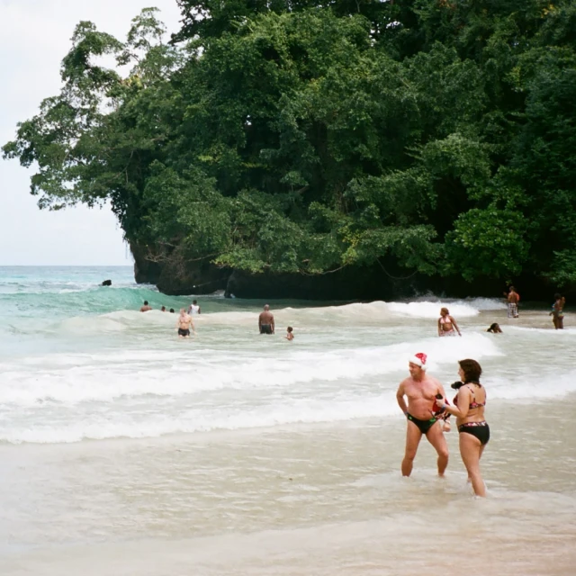 People in swimwear wade in the ocean surf near a beach with dense tropical foliage. One man wears a Santa hat in the bright daylight.