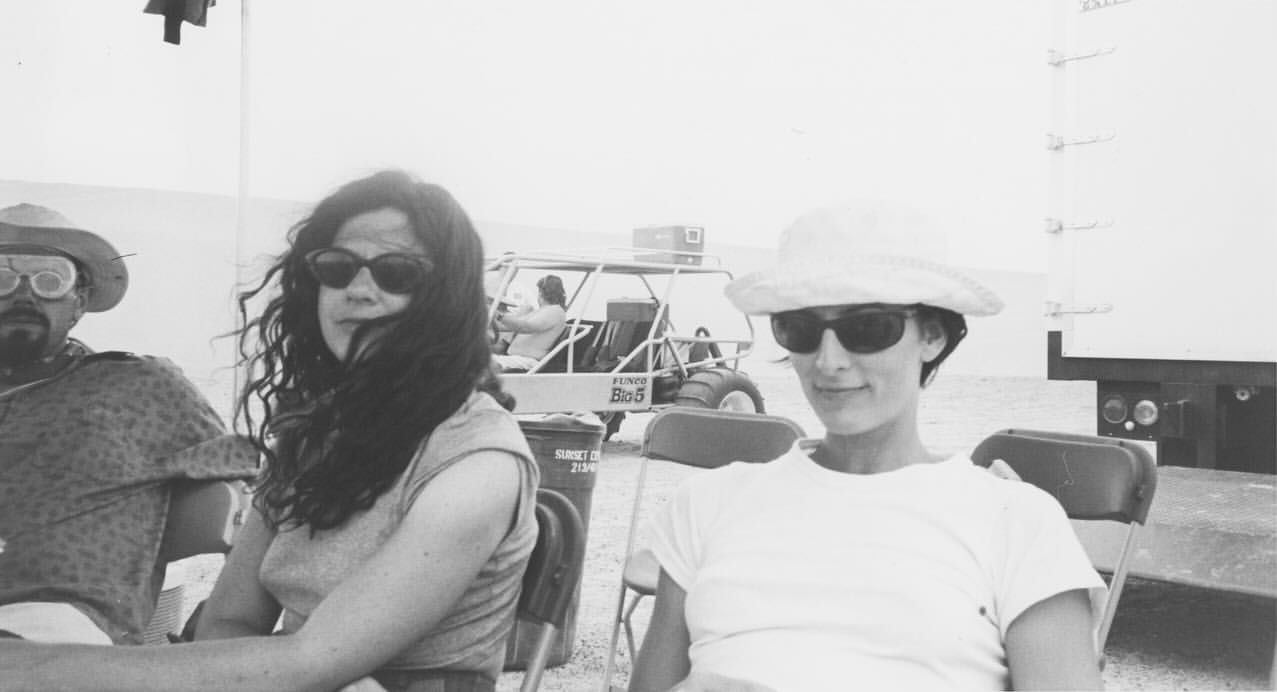 Black and white photo features a group of people outdoors with a dune buggy and truck. Two women and a man are seated and smiling.