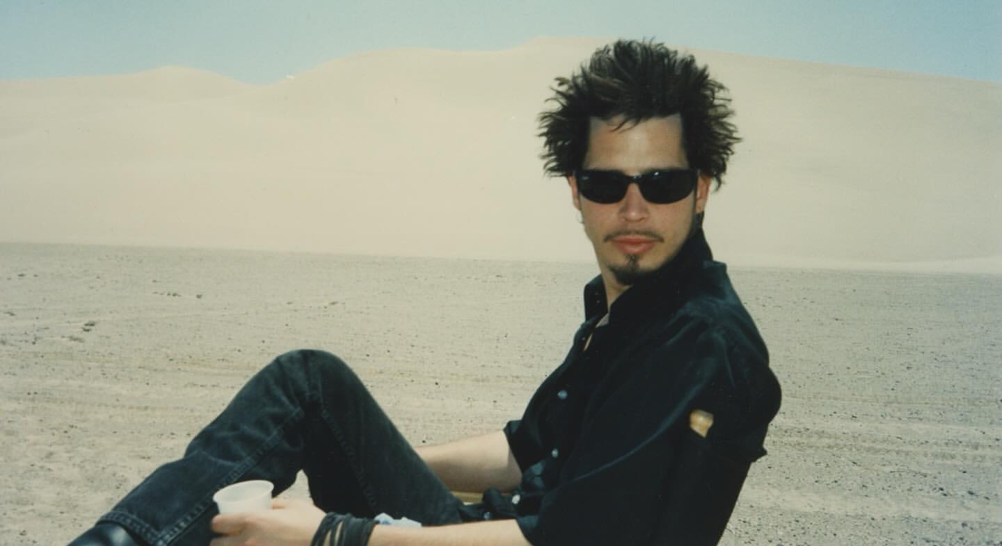 Adult male with sunglasses sits in the desert with sand dunes in background, wearing a black shirt and jeans, holding a cup.