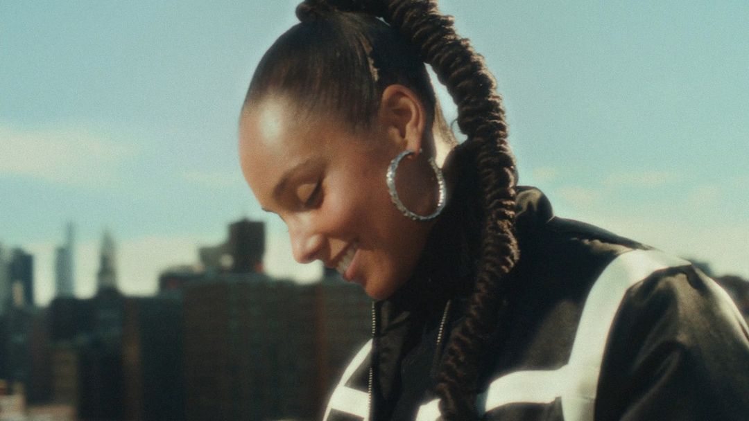 Alicia Keys smiling with a high braided ponytail and hoop earrings outdoors against a city skyline by Cheryl Dunn
