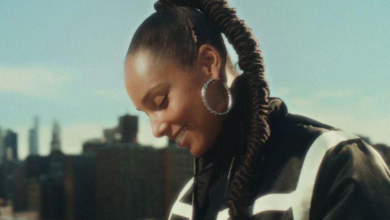 Alicia Keys smiling with a high braided ponytail and hoop earrings outdoors against a city skyline by Cheryl Dunn