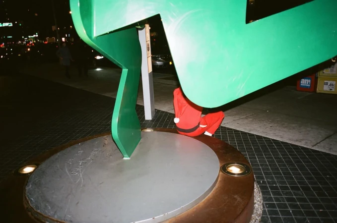 Low-angle shot of a person in a Santa suit crouched beneath a large green arrow sculpture at night, set against an urban backdrop.