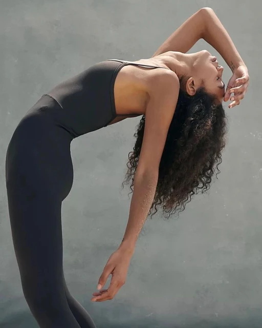A Black woman in a dark bodysuit arches backward with her head tilted back and her arms at her sides in a fitness pose on grey backdrop.
