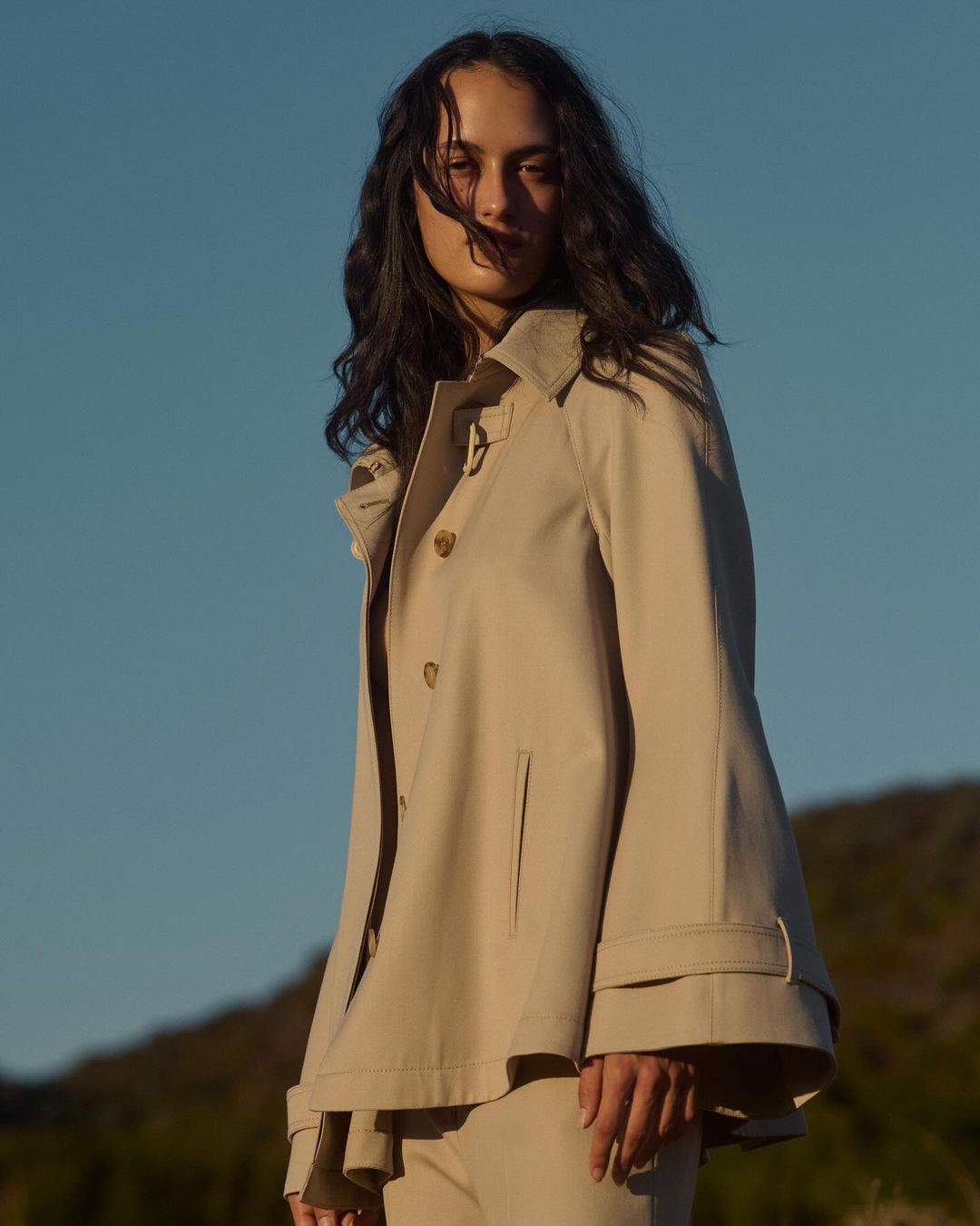 Woman in a beige trench coat poses outdoors against a clear blue sky. The low angle and bright daylight highlight the clothing.