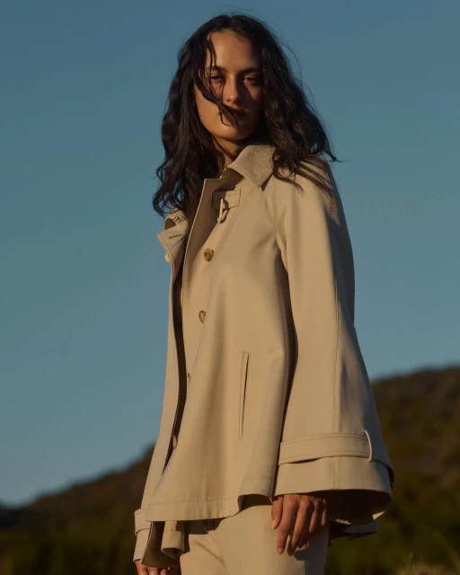 Woman in a beige trench coat poses outdoors against a clear blue sky. The low angle and bright daylight highlight the clothing.