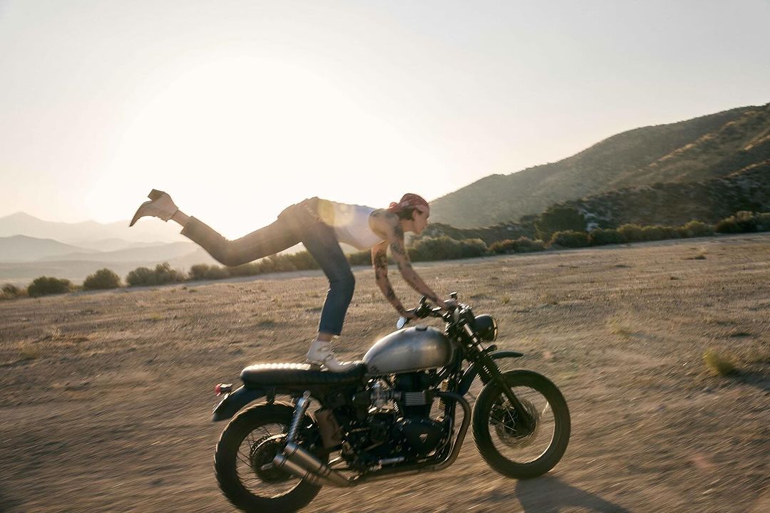 Woman stands on a vintage motorcycle in the desert. The background shows desert landscape with mountains and a bright sunny sky.