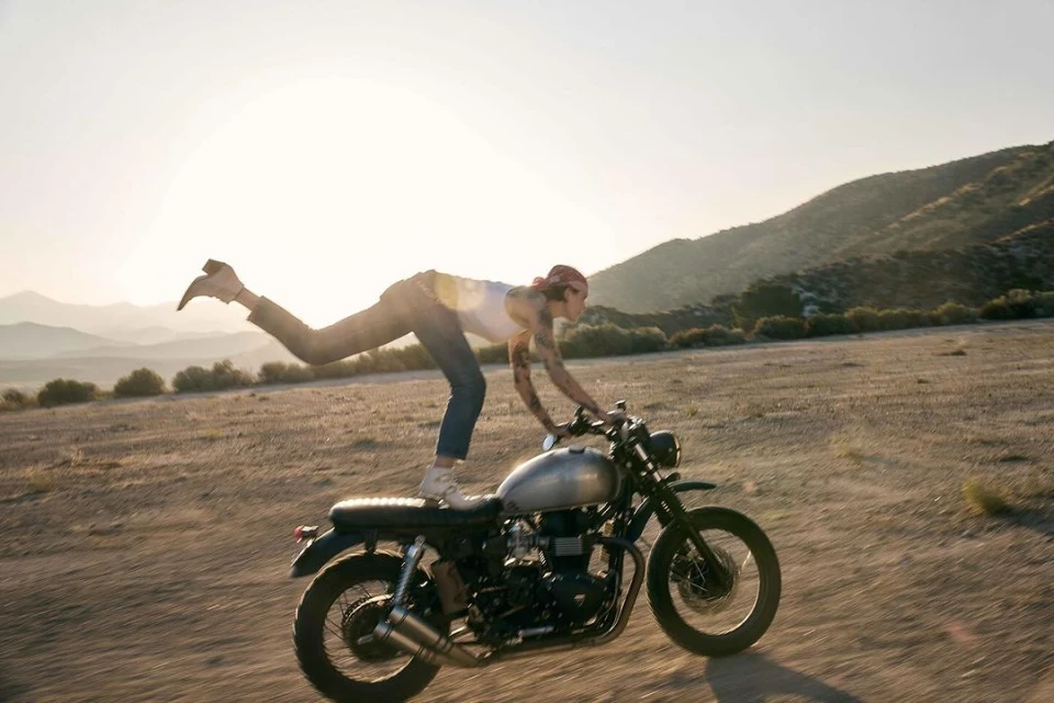 Woman stands on a vintage motorcycle in the desert. The background shows desert landscape with mountains and a bright sunny sky.