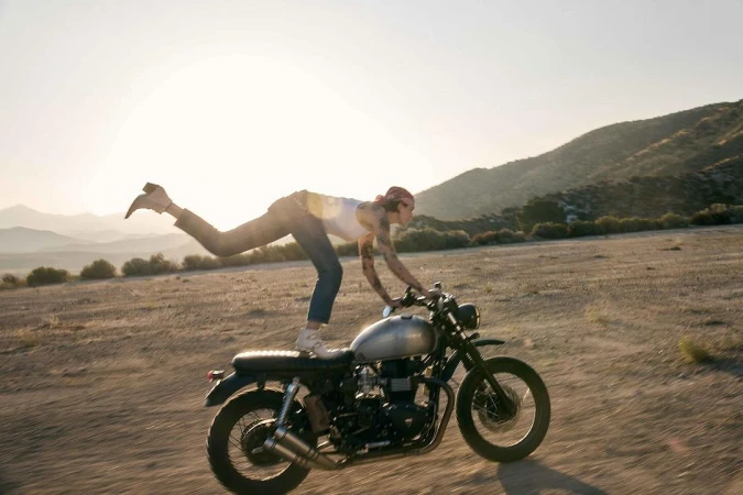 Woman stands on a vintage motorcycle in the desert. The background shows desert landscape with mountains and a bright sunny sky.