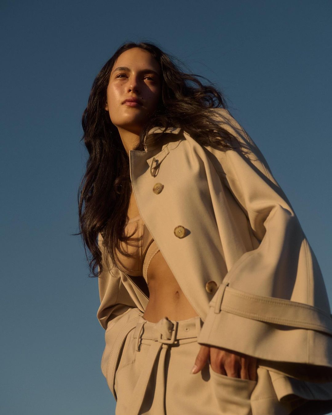 Woman in a tan trench coat and pants, looking up against a clear blue sky, outdoor fashion shot, eye-level view.