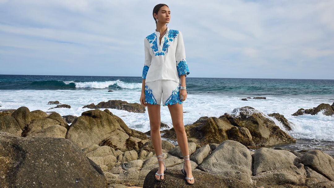 Model standing on coastal rocks wearing a white matching set with blue floral embroidery against an ocean background