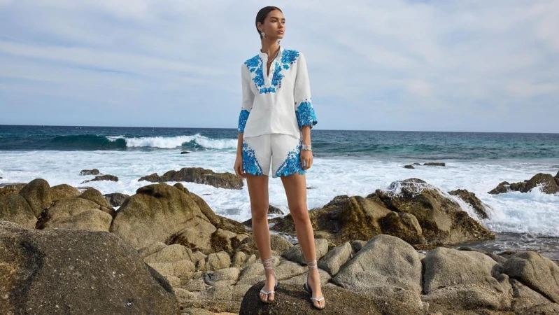 Model standing on coastal rocks wearing a white matching set with blue floral embroidery against an ocean background