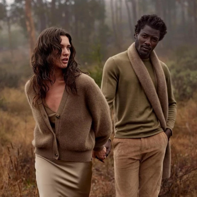 A fashionable couple, a brunette woman in a cardigan and a Black man with a scarf, stands in an out-of-focus wooded area.