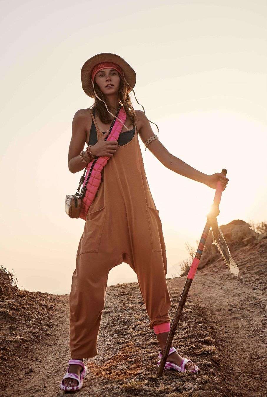 A woman stands on a sandy path wearing a jumpsuit, hat, and sandals. She is holding a wooden walking stick in the desert.