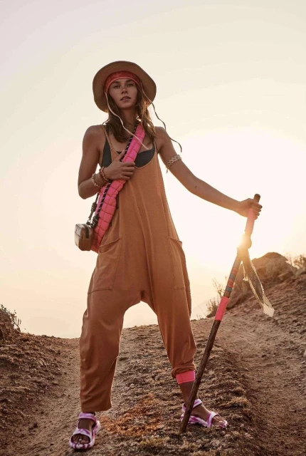 A woman stands on a sandy path wearing a jumpsuit, hat, and sandals. She is holding a wooden walking stick in the desert. 