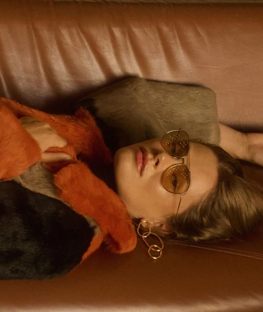 Woman in sunglasses with orange fur coat lying on a peach-colored couch with gold earrings.
