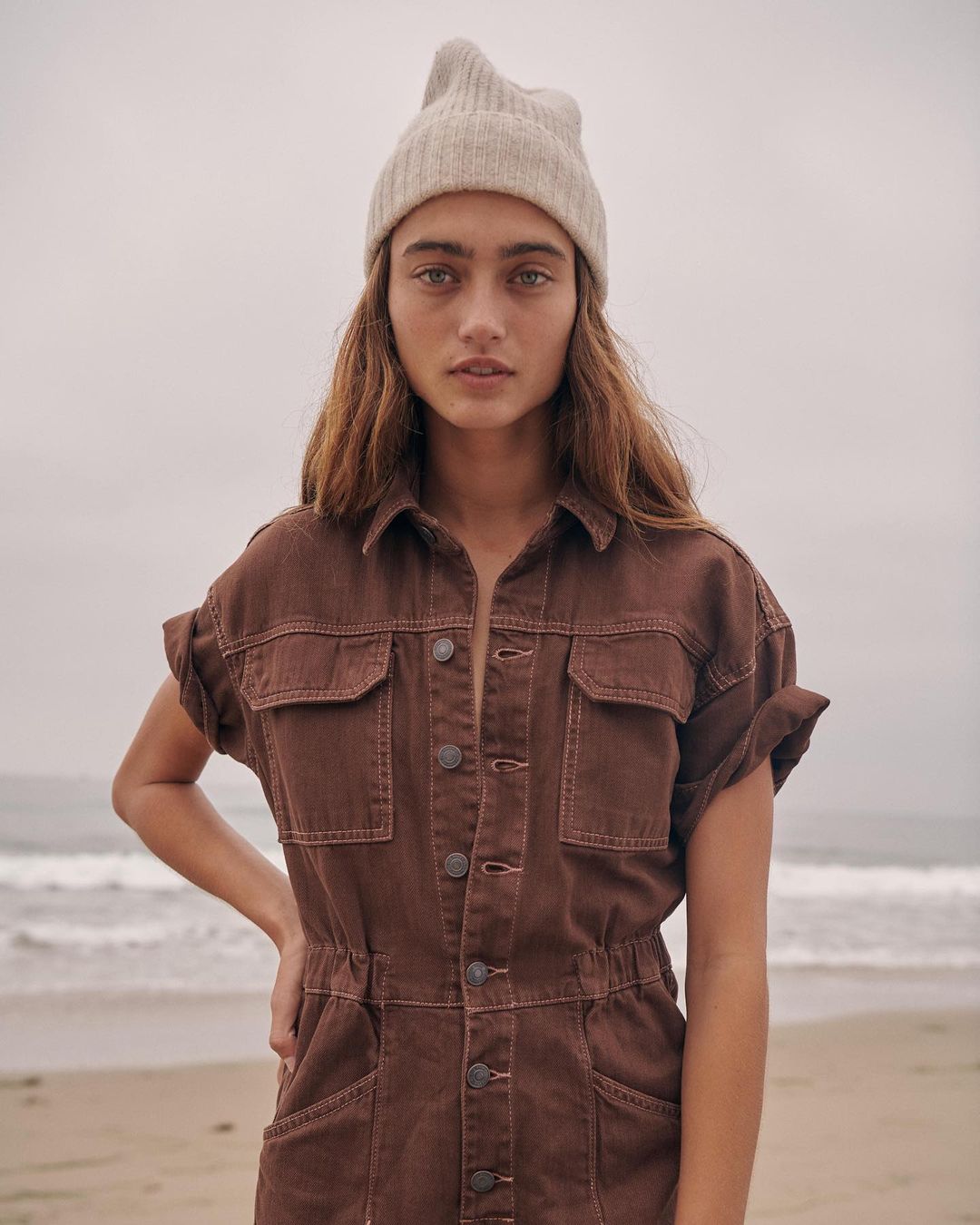 Portrait of a woman in a brown jumpsuit and beanie on a beach with muted tones, featuring soft, diffused lighting and a serene atmosphere.