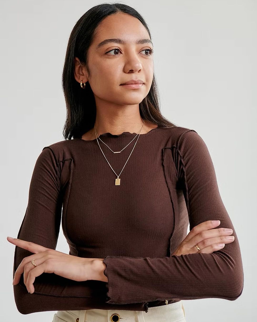 A woman wearing a brown ribbed long-sleeve top and layered gold necklaces against a plain light background