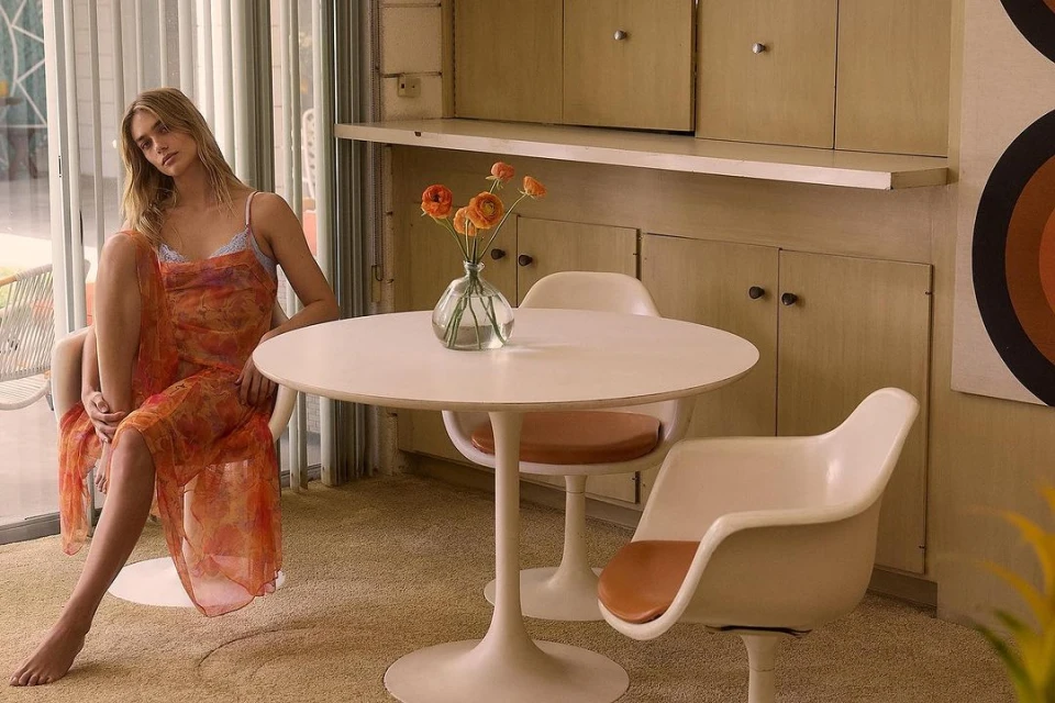 A barefoot, adult female model poses indoors while sitting next to a table with flowers, wearing a floral dress in a casual indoor setting.
